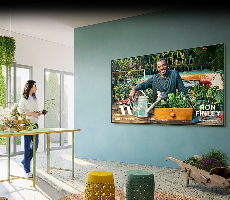 Hay un TV grande en la pared y muestra una clase de jardinería de ?Master Class?. Una mujer está sentada en una mesa junto a un TV con macetas y tijeras de podar, siguiendo una clase de jardinería.