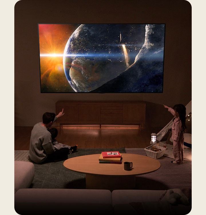 Une famille assise par terre dans une pièce à la lumière tamisée à c?té d’une petite table, qui regarde un téléviseur LG installé sur le mur montrant la Terre depuis l’espace.