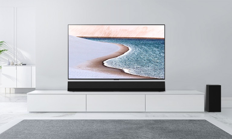 Un téléviseur est monté sur un mur gris clair. La barre de son LG se trouve en dessous, sur un meuble blanc. Le téléviseur montre une plage.