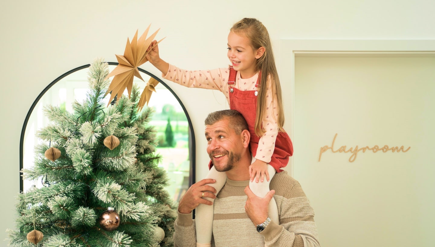 Un père porte sa fille souriante sur ses épaules pendant qu’elle décore le sapin de No?l. Une porte marquée 'salle de jeux' est visible à proximité.