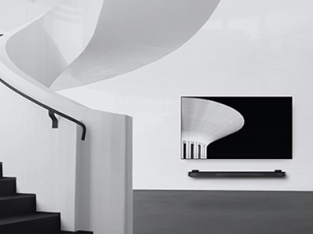 a black and white image of lg signature oled tv which is hung on the wall and to the left there is a spiral staircase