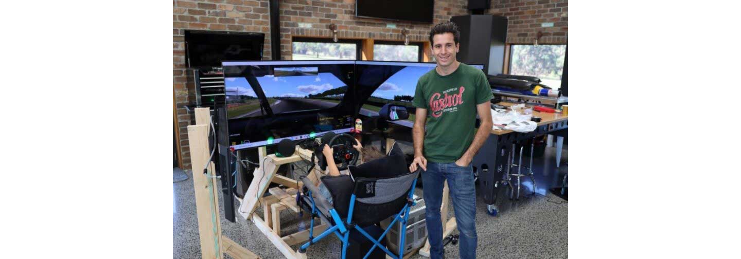 Rick Kelly poses and in front of his new racing simulator as his son uses it behind him