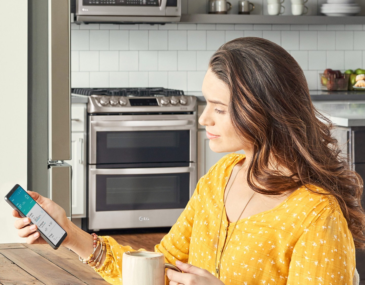 Woman sitting in her kitchen looking at a phone displaying the ThinQ? app screen showing the device status of the washer, refrigerator and range