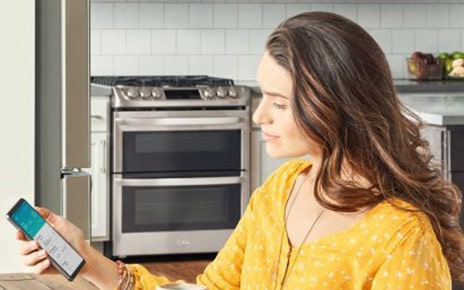Woman sitting in her kitchen looking at a phone displaying the ThinQ? app screen showing the device status of the washer, refrigerator and range