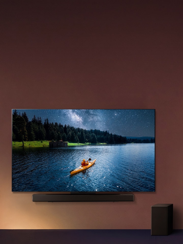LG TV dan Soundbar dipasang di dinding dan subwoofer di lantai sebelah kanan. Di TV, seseorang yang dengan kayak di danau ditampilkan, dan bayangan halus mengalir di dinding.