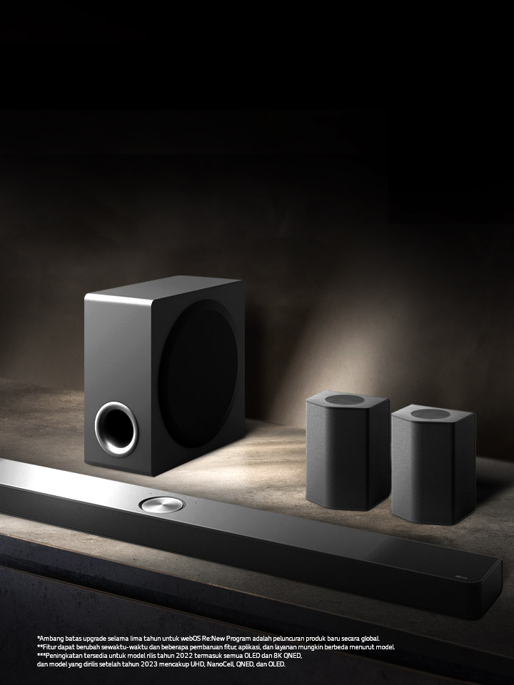 LG Soundbar, speaker belakang, dan subwoofer ditempatkan dalam perpektif bersiku pada rak kayu coklat di dalam ruangan hitam, tertutup dalam kegelapan dengan hanya ada cahaya yang menerangi sound system.