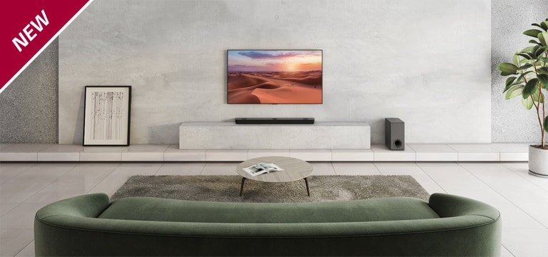 Il televisore è appeso al pilastro bianco al centro del soggiorno. Sotto, la Sound Bar LG è collocata su una mensola bianca. Sul pavimento in legno, proprio sotto la sound bar, troviamo un subwoofer wireless nero. Dietro il divano verde, due diffusori posteriori sono posizionati sul tavolino da caffè sia a sinistra che a destra. Il testo NEW (NOVITà) viene visualizzato nell’angolo superiore sinistro.