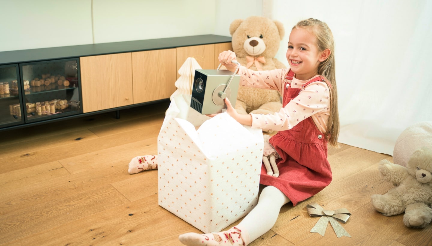 Una giovane ragazza sorridente apre un regalo di Natale e trova un proiettore LG CineBeam. è seduta su un pavimento in legno.