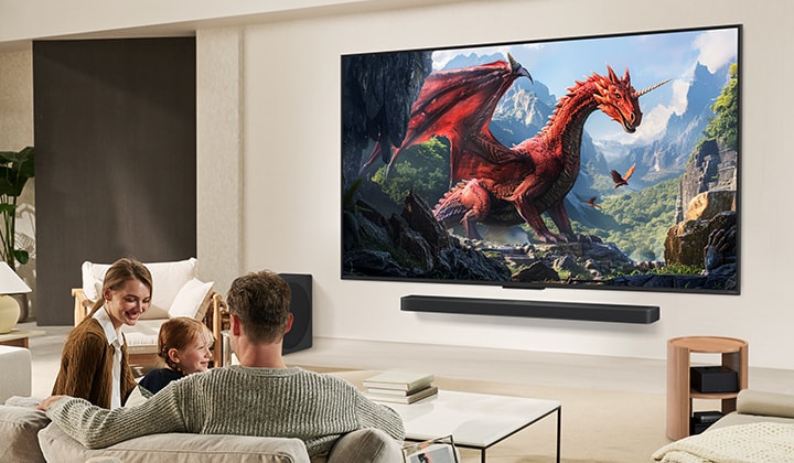 Una familia se sienta en un sofá frente a un televisor LG QNED montado en una pared sobre una barra de sonido LG con una ni?a se?alando una pantalla que muestra dos delfines.