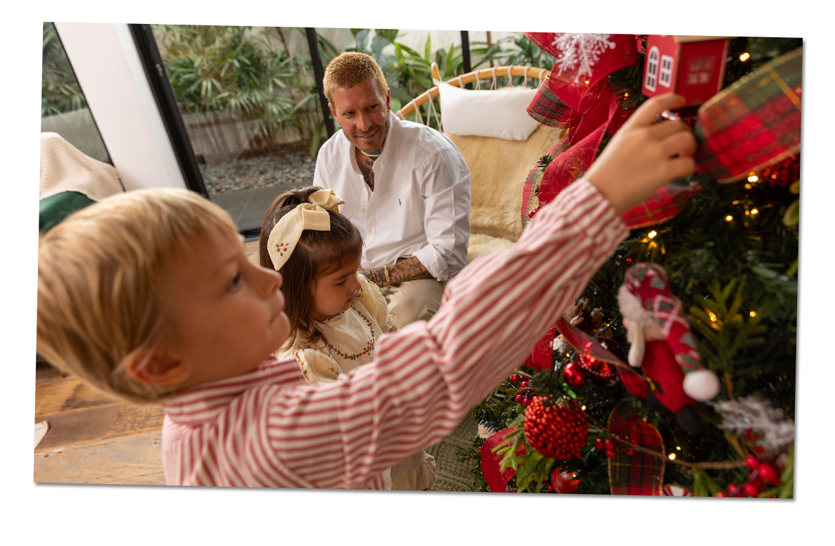 Un padre y sus hijos se divierten decorando un árbol de Navidad. El ni?o sostiene un adorno para colocarlo en el árbol.