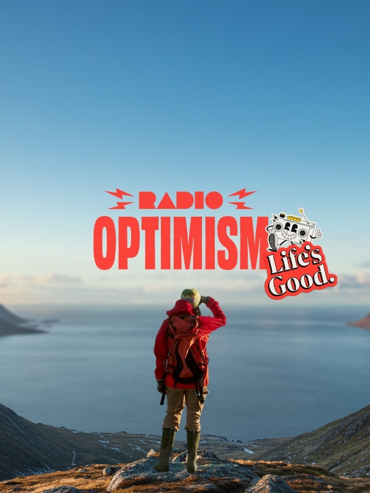 Una mujer con una chaqueta de senderismo roja está de pie en un lugar donde se puede ver el vasto océano y monta?as cubiertas de nieve. La imagen también presenta el logotipo del eslogan "Radio Optimism" y el logotipo de "Life's Good." junto a la mujer.