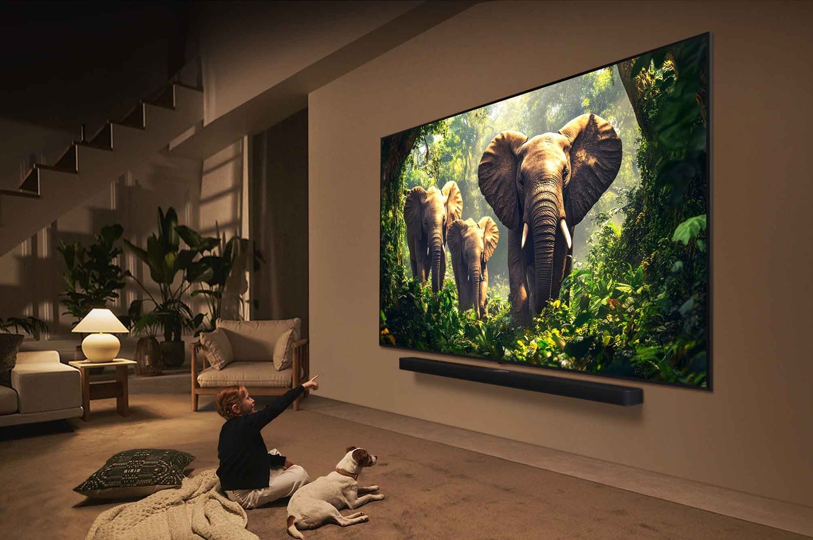 A girl and a dog are seated in front of a LG QNED TV mounted on a wall depicting three elephants walking outwards above a LG Soundbar. The title talks about how every action is more thrilling on a massive screen.