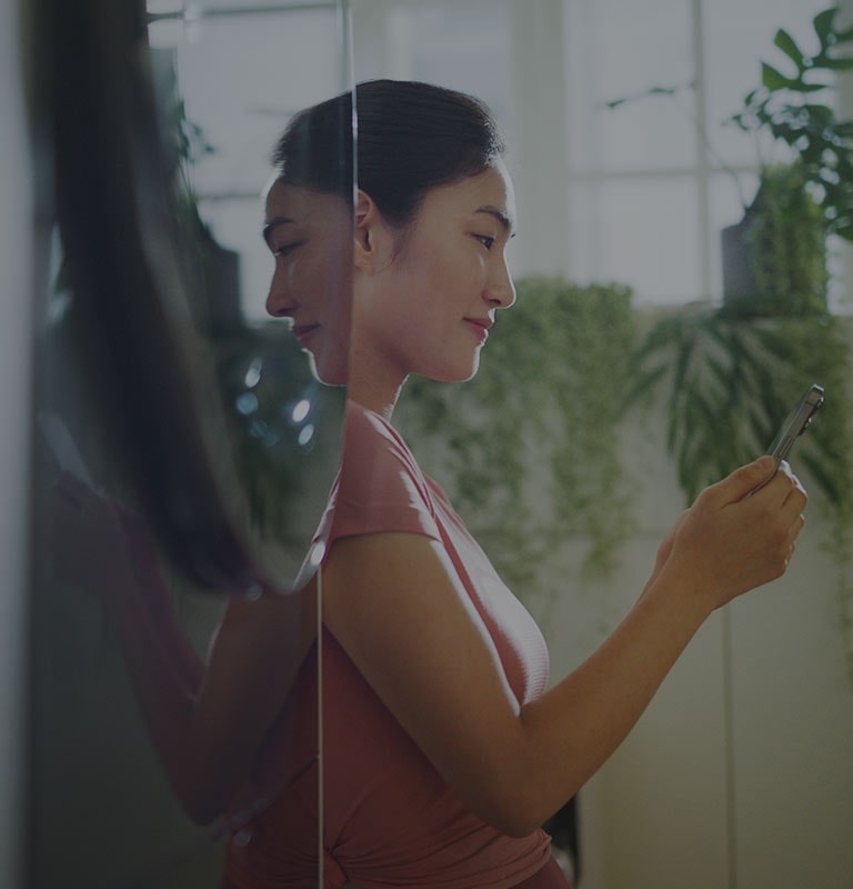 La imagen muestra a una mujer de pie junto a una torre de lavado LG WashTower?, mirando su teléfono mientras sonríe.