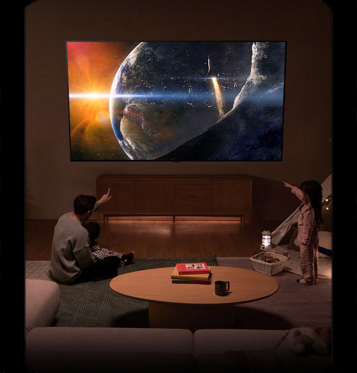 Una familia sentada en el suelo de una sala de estar poco iluminada frente a una mesa peque?a mirando un LG TV montado en la pared que muestra la Tierra desde el espacio.