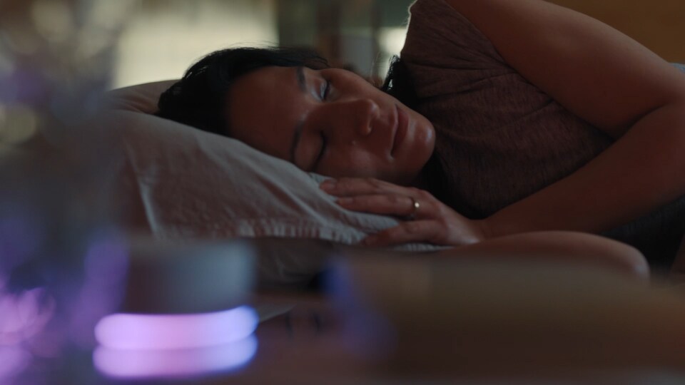 A woman lies on her side, sleeping in bed. In front of her, the LG ThinQ ON device is glowing. 
