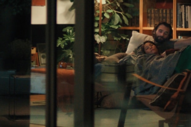 A scene where the camera captures a man and a woman lying together on the living room sofa from a distance.