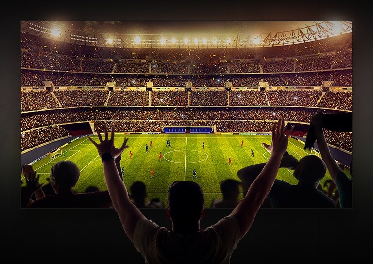 Quatro pessoas sentadas num sofá a ver TV num ecr? grande com imagem de um estádio de futebol cheio de pessoas a assistir.