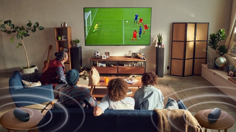 Cinco pessoas sentadas no sofá a ver uma TV de grande ecr? com um jogo de futebol com colunas Bluetooth a fazer o som surround.