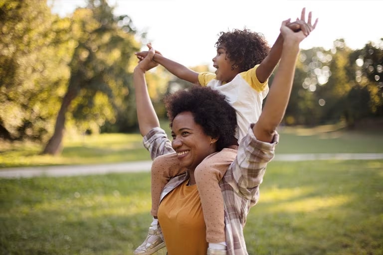  Afroamerikansk mamma och dotter som st?r i en park. Liten flicka p? mammas axlar