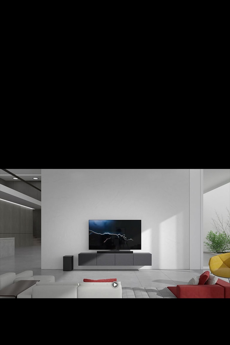 Soundbar, oturma odas?nda bir TV ile gri dolaba yerle?tirilmi?tir. Sol taraftaki zemine kablosuz bir siyah subwoofer yerle?tirilmi?tir ve resmin sa? taraf?ndan güne? ????? gelir. Beyaz ve k?rm?z? renkli uzun bir kanepe, TV'ye ve soundbar'a bakacak ?ekilde yerle?tirilmi?tir.