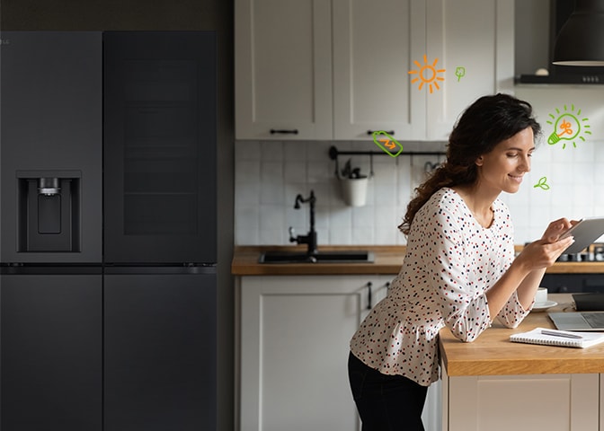 La femme postée devant le réfrigérateur est en train de sourire en regardant le panneau, et autour d’elle, des ampoules électriques et des ic?nes représentant la nature illustrent l’efficacité énergétique.