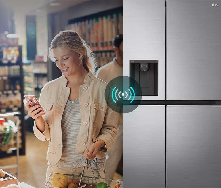 A woman at a grocery store uses her smartphone while standing next to an LG refrigerator with Wi-Fi connectivity.