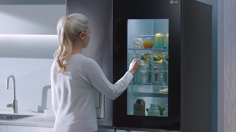 Woman with ponytail retrieves drink from illuminated LG fridge with transparent door in modern kitchen.