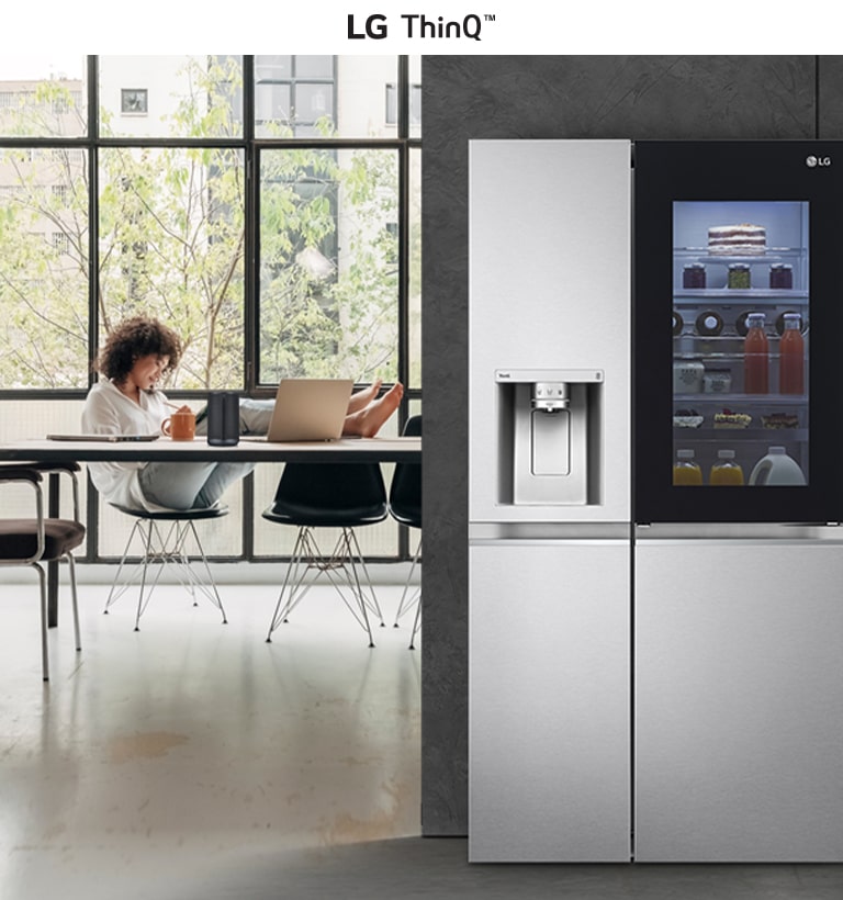 LG ThinQ smart fridge with glass panel next to a modern kitchen workspace with large windows.