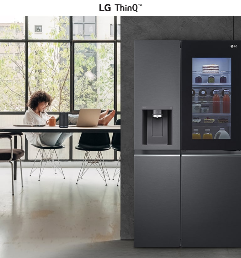 Modern kitchen with an LG ThinQ refrigerator on the right and a person using a laptop by a large window on the left.