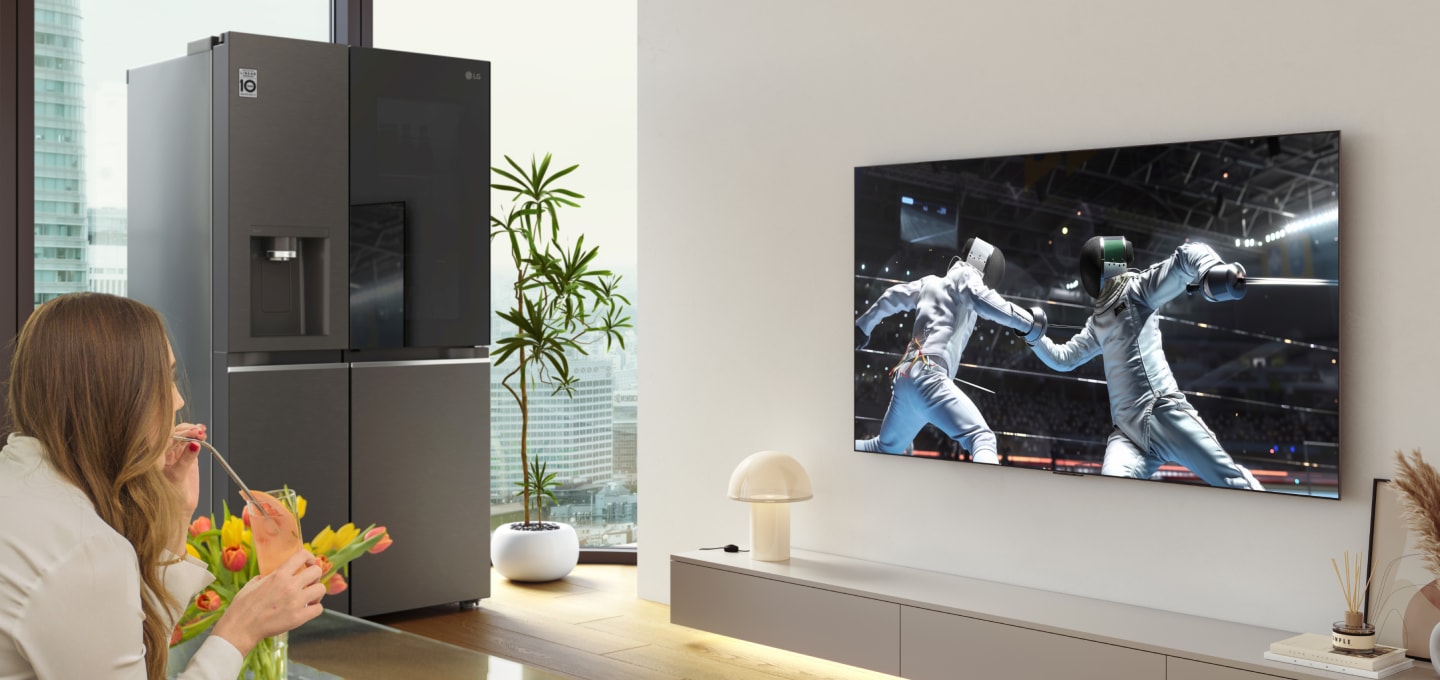 A woman watching a sports game on an LG OLED TV in the living room, holding a mocktail, with an LG InstaView refrigerator next to the TV wall.