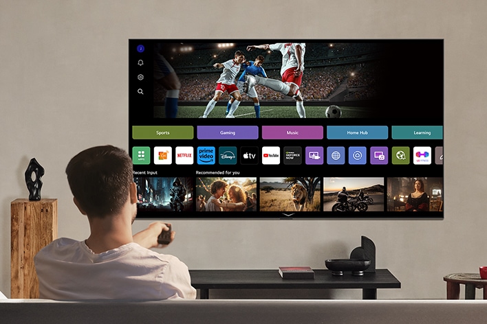 Ng??i trên gh? dài ?ang c?m ?i?u khi?n t? xa. LG AI TV treo t??ng, trên màn hình là LG webOS.