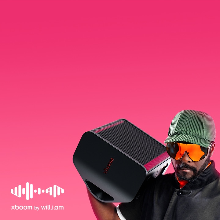 will.i.am m?c trang ph?c ?en và ?eo kính ram, ?ang vác chi?c XBOOM Stage 301 trên vai.