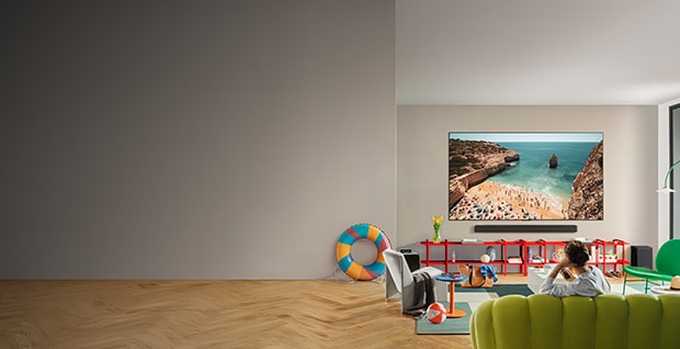Person som sitter i en limef?rgad soffa, matta och f?t?lj med somriga f?rem?l runt omkring sig, en badring, en strandboll och en TV p? v?ggen med en bild av en strand vid Medelhavet.