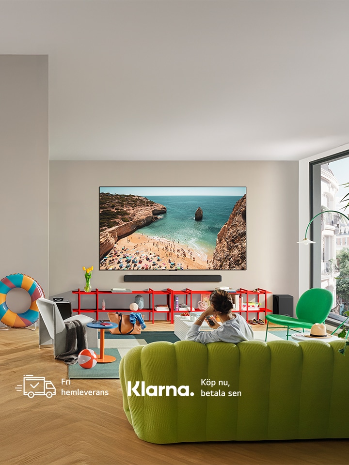 Person som sitter i en limef?rgad soffa, matta och f?t?lj med somriga f?rem?l runt omkring sig, en badring, en strandboll och en TV p? v?ggen med en bild av en strand vid Medelhavet.