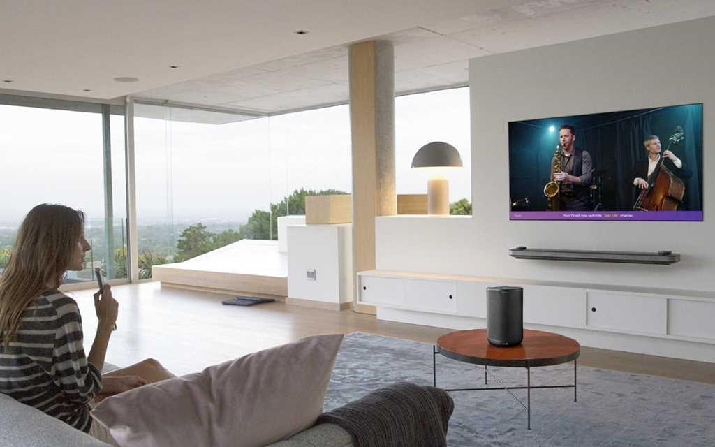 ?ena sedí na pohovce v obývacím pokoji a pomocí LG dálkového ovládání mluví na její LG OLED Wallpaper TV. 