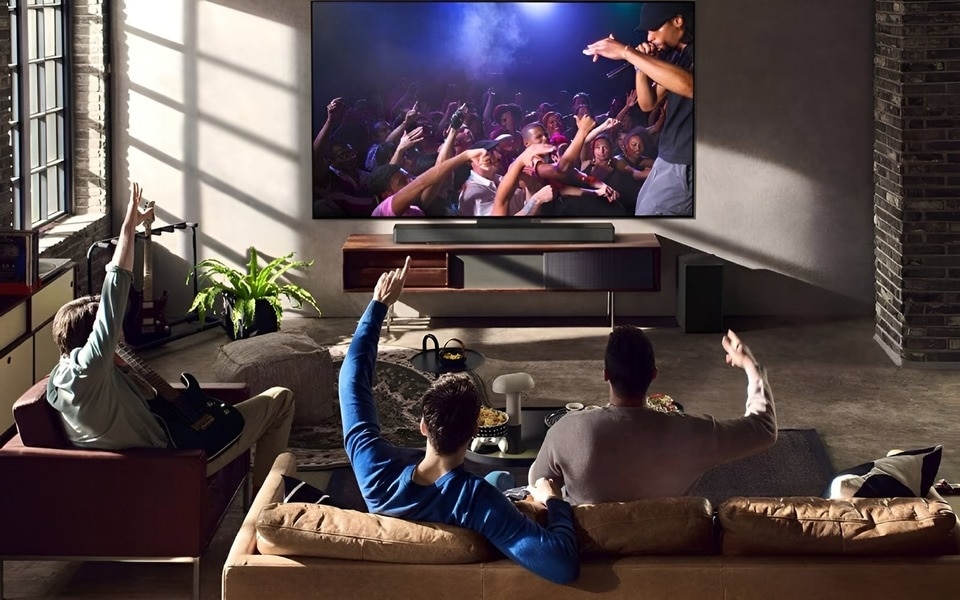 Eine Gruppe von Freunden in einem Loft-Wohnzimmer, die sich ein Live-Konzert auf einem großen Fernsehbildschirm mit preiswerter LG Soundbar und Subwoofer ansehen. Die Atmosphäre ist gemütlich und lebendig und veranschaulicht das Surroundsound-Erlebnis.