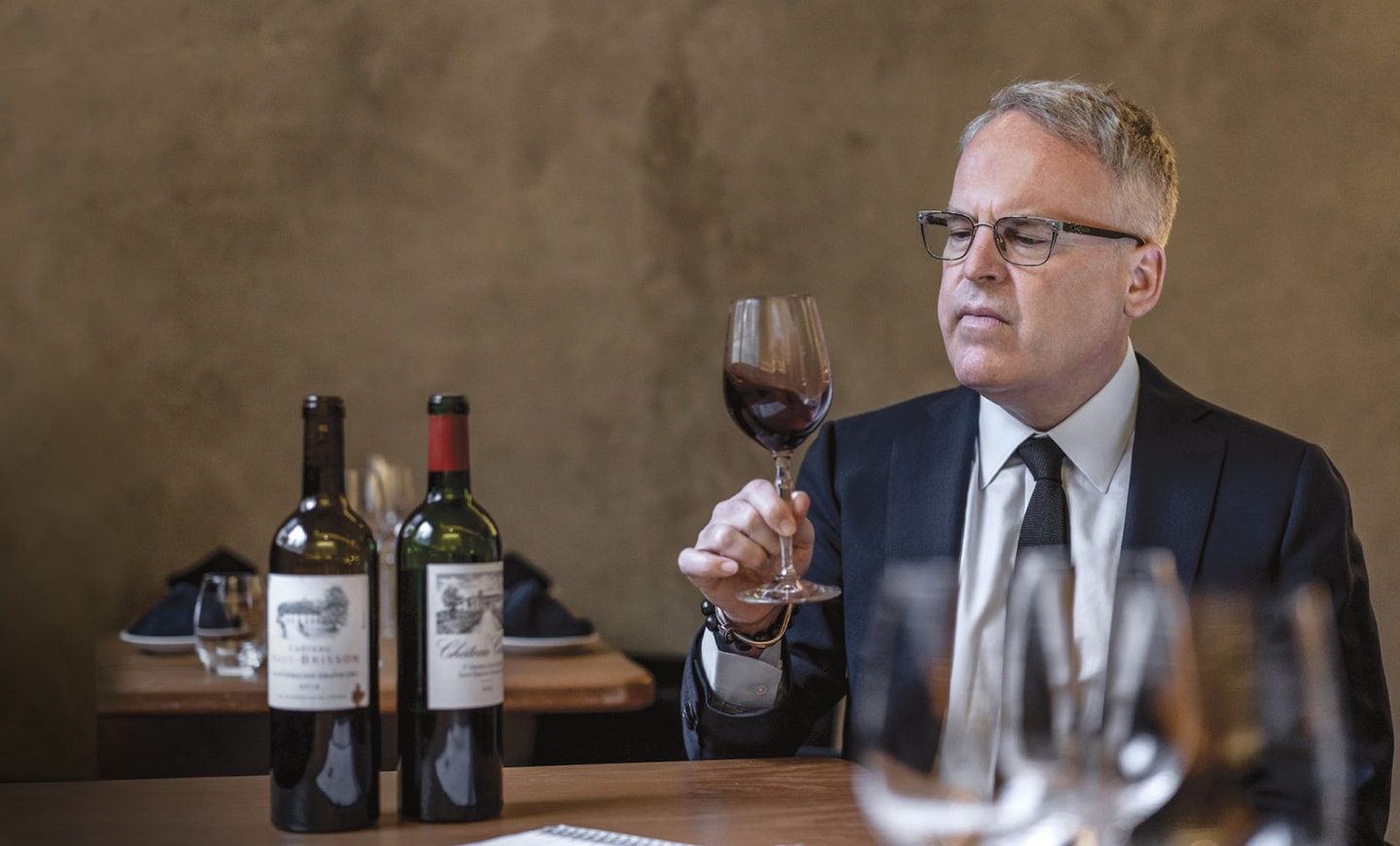 James Suckling h?lt ein Glas Wein, w?hrend vor ihm zwei Weinflaschen auf dem Tisch stehen.
