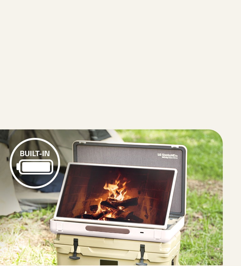 El LG StanbyME Go se coloca frente a una tienda de campa?a y la pantalla muestra uno de los temas relajantes: una chimenea. En la esquina superior izquierda, se muestra el ícono de la batería incorporada.