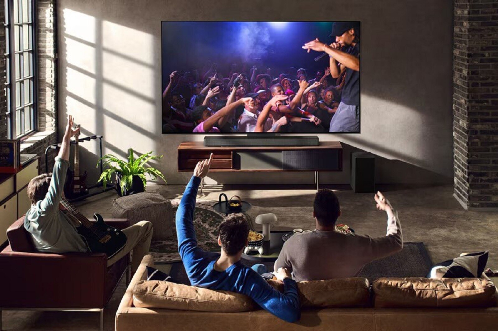 Un salon de style loft dans lequel un groupe d’amis regarde un concert en direct sur un grand téléviseur équipé d’une barre de son LG et d’un caisson de basses. L’ambiance est chaleureuse et dynamique, mettant en valeur la qualité du son surround. 