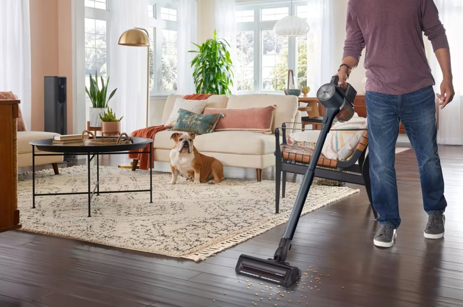 Personne utilisant un aspirateur balai sans fil sur un sol en bois dans un salon moderne avec un bulldog assis sur un tapis.