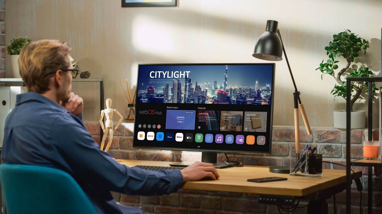 une personne assise à un bureau utilise un Smart Monitor LG affichant une interface LG webOS avec plusieurs applications. L’espace de travail comprend une lampe, des plantes et une décoration moderne avec un mur de briques, créant un environnement de travail confortable.