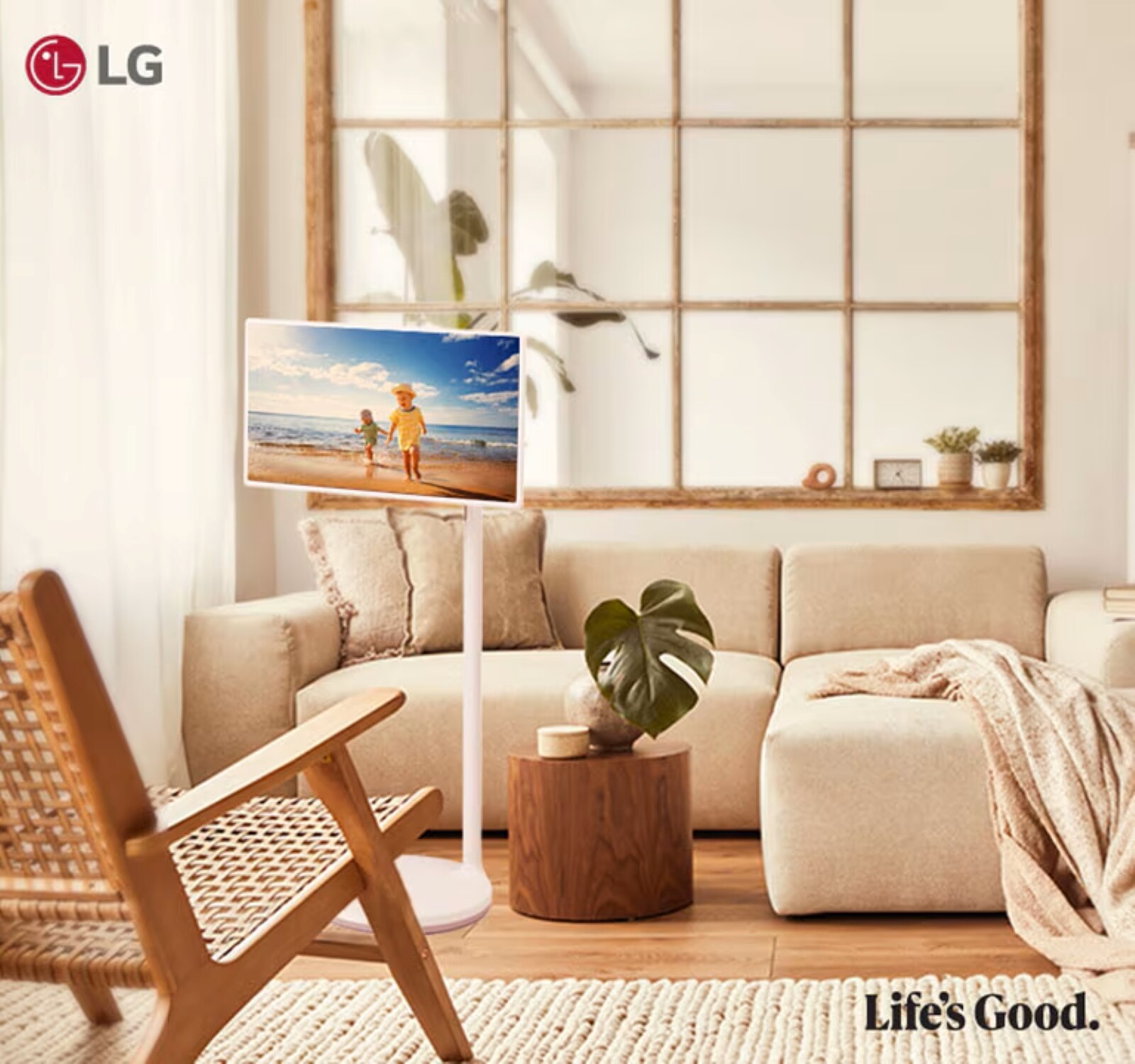 Un salon lumineux et cosy avec des tons neutres, comprenant une grande fenêtre, un canapé beige confortable, une chaise en bois et un écran numérique affichant une scène de plage. Le logo LG et le slogan "Life's Good" sont visibles dans le coin.