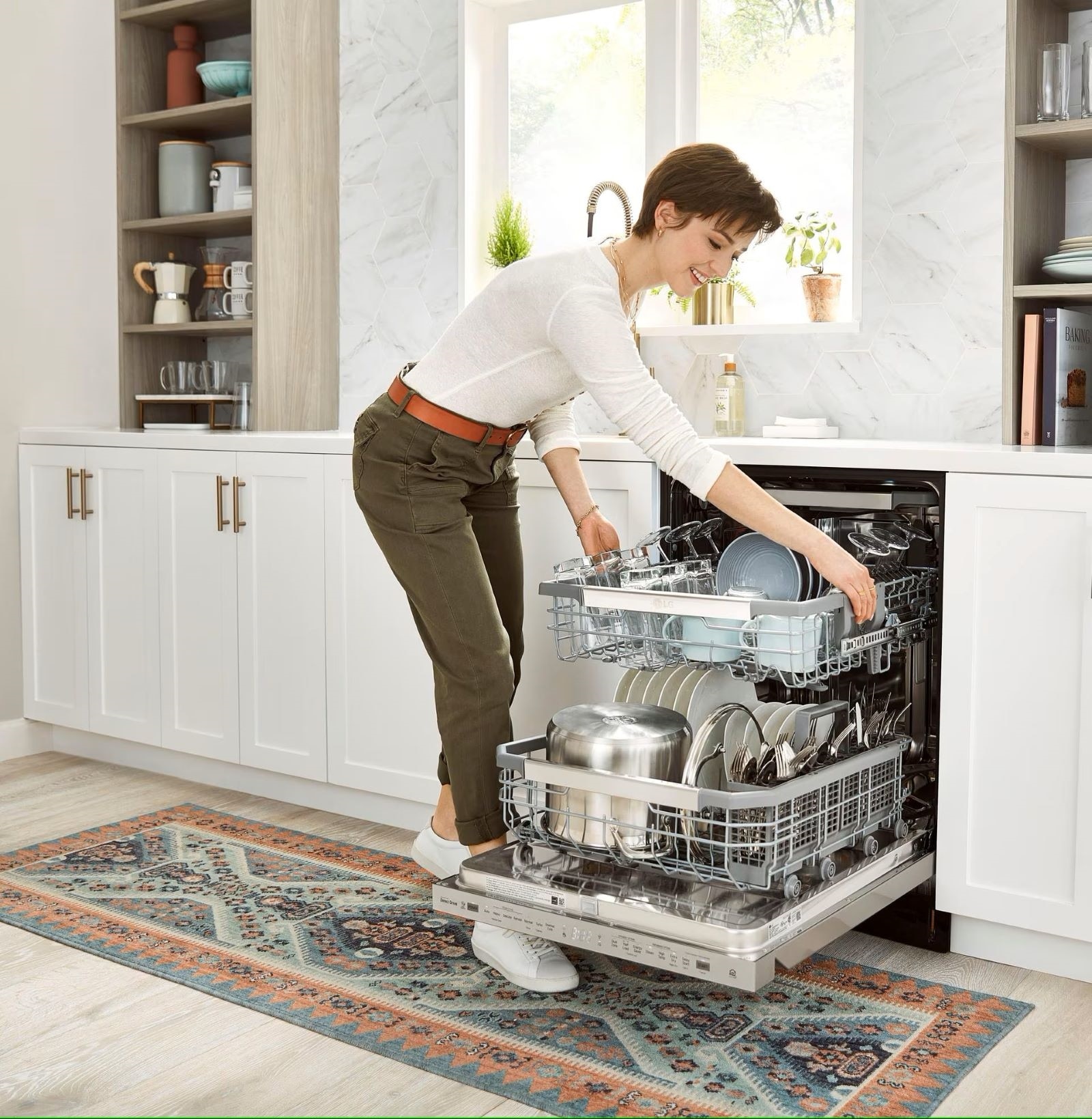 Une femme est en train de charger un lave-vaisselle dans une cuisine moderne composée de placards blancs et d’un tapis à motifs au sol. Le lave-vaisselle est entièrement ouvert et elle dépose un plat sur le panier supérieur.