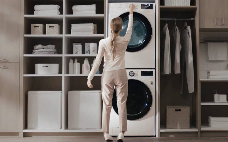 Une femme ajuste les réglages d’un lave-linge et sèche-linge blancs empilés dans un espace buanderie bien organisé, avec des étagères contenant des produits de lessive soigneusement rangés et des vêtements pliés.