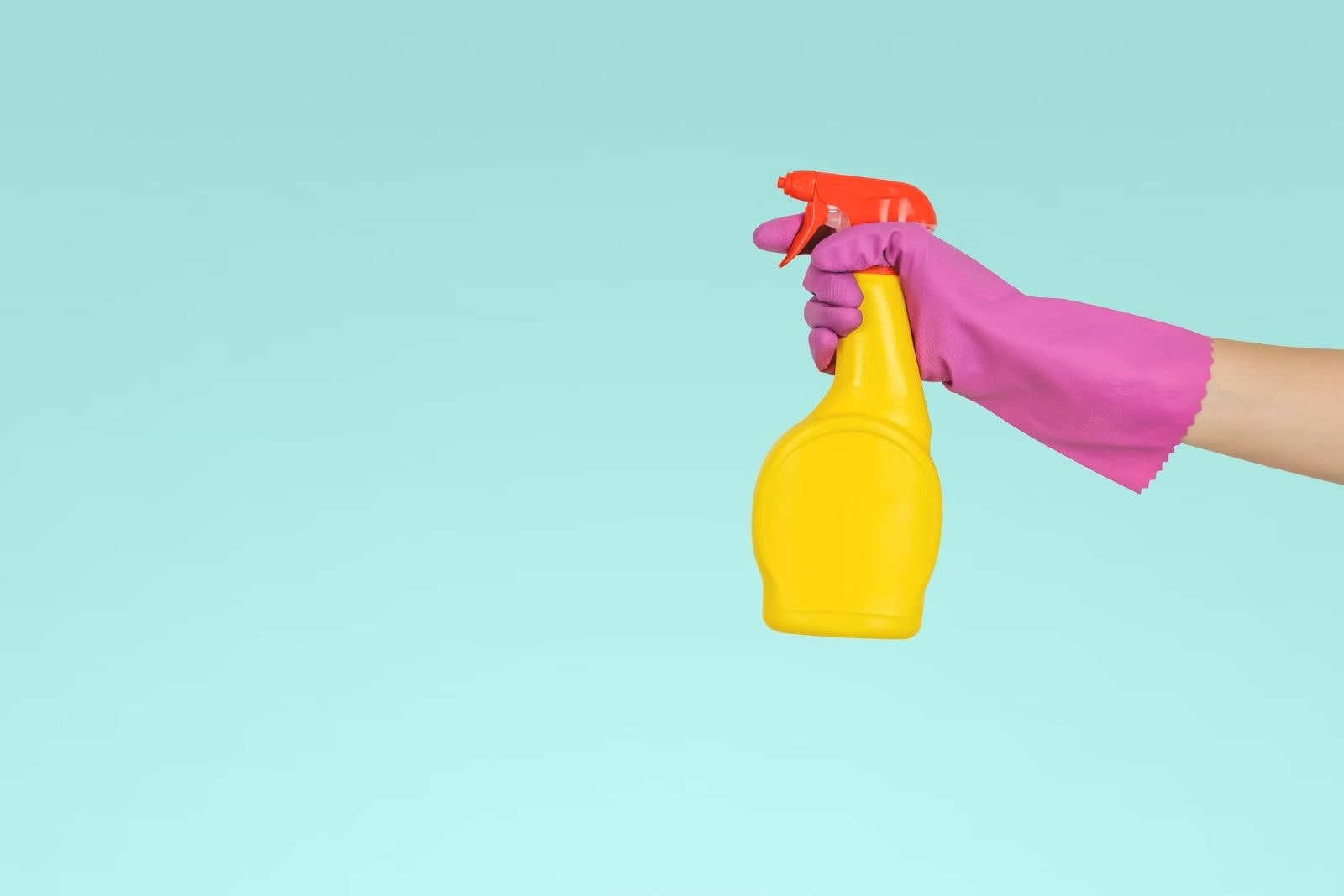 Main portant un gant rose tenant un vaporisateur jaune avec un bouchon rouge, utilisé pour nettoyer un frigo et enlever les mauvaises odeurs, sur un fond bleu clair.