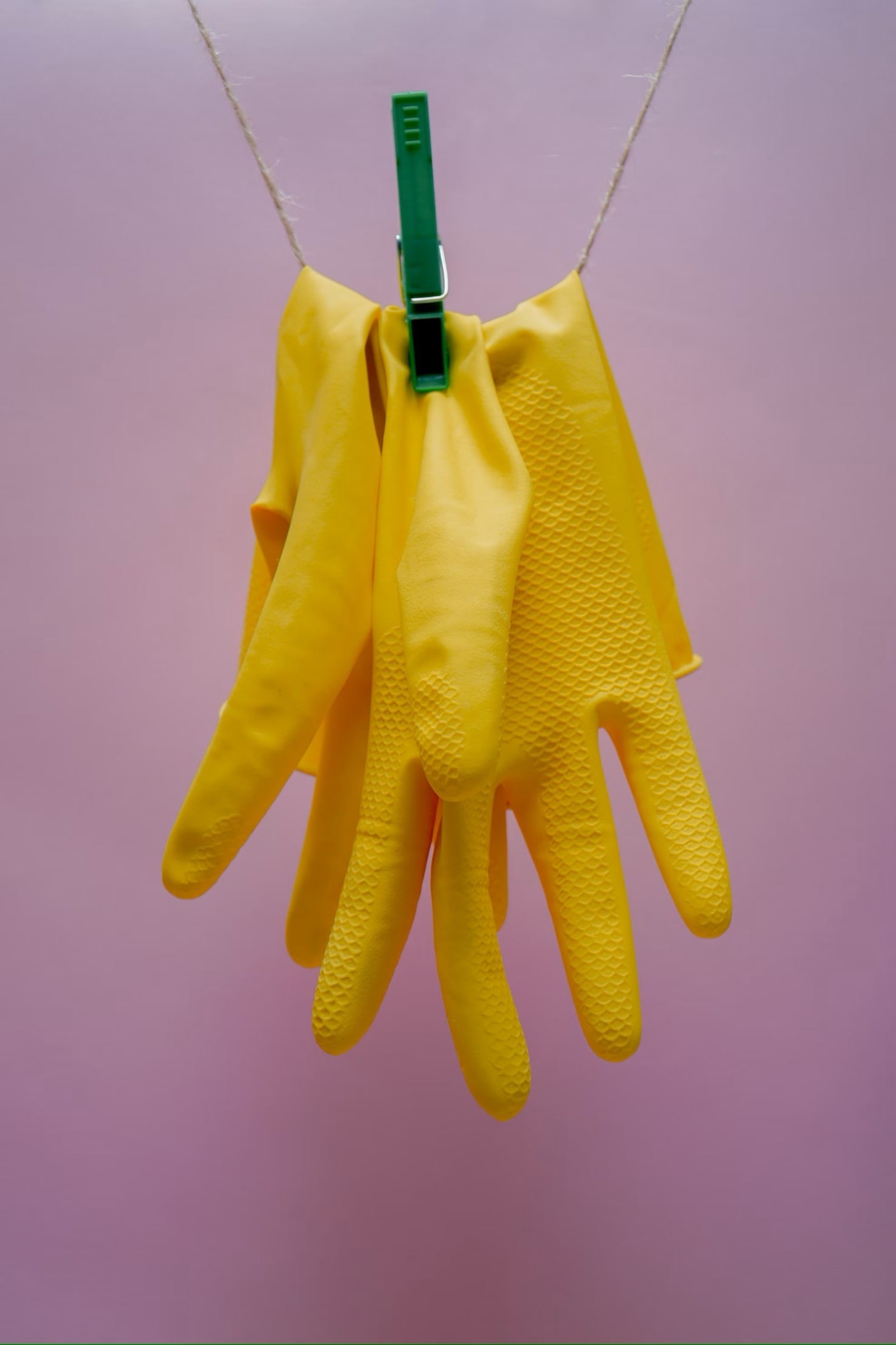 Gants de nettoyage jaunes suspendus à une corde à linge avec une pince à linge verte sur un fond rose, symbolisant la propreté et les techniques de nettoyage pour réfrigérateurs en inox, y compris l'élimination des odeurs et le nettoyage des trous de drainage.