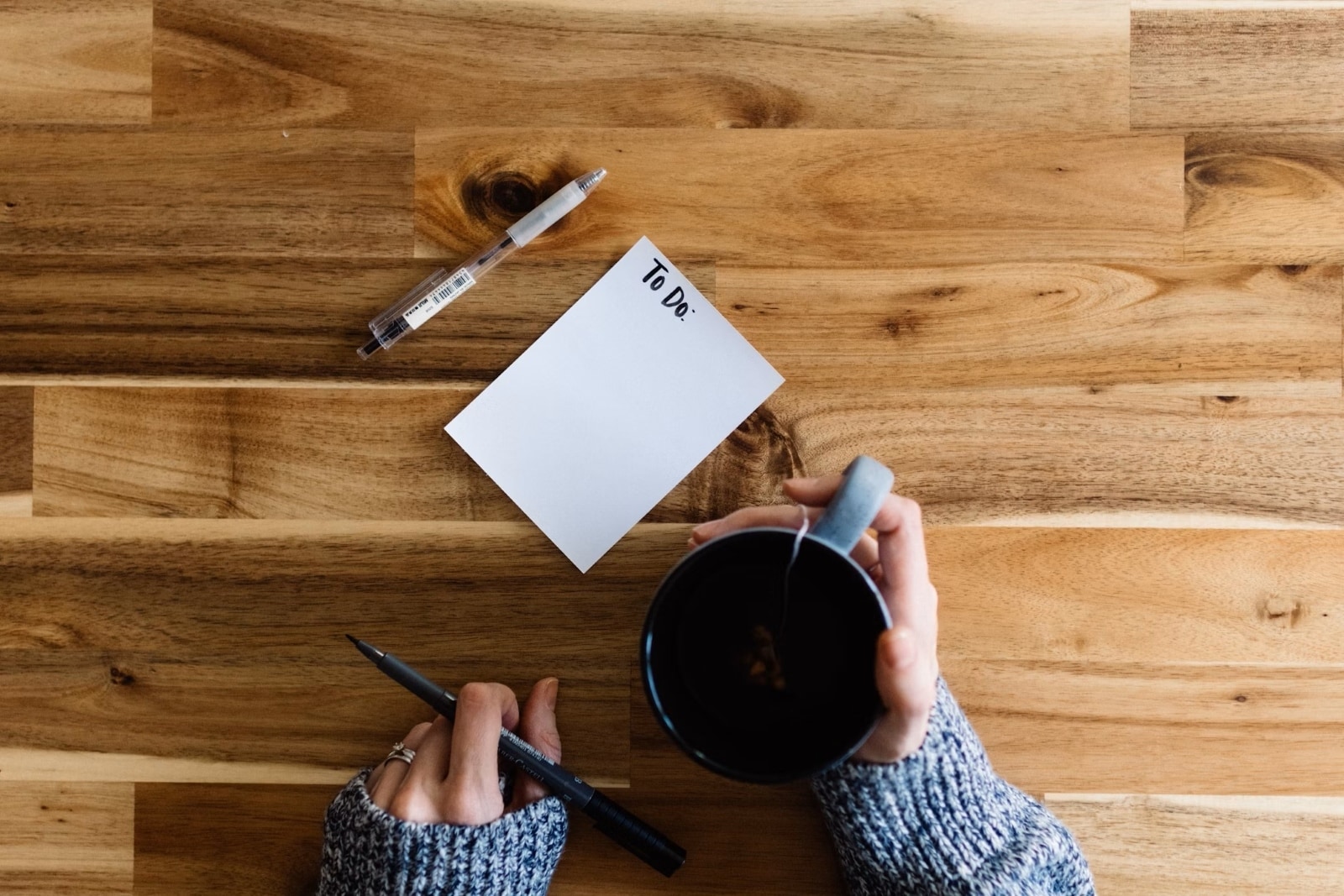 Personne tenant une tasse de café et un stylo près d'une liste de tâches "To Do" sur une table en bois.