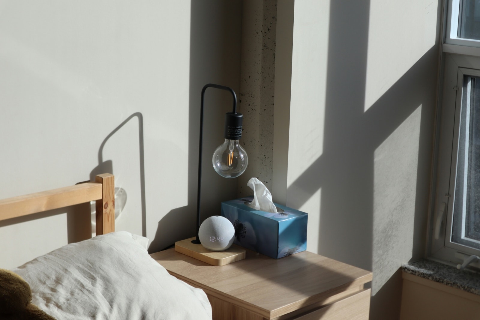 Une table de nuit avec un haut-parleur intelligent dans une chambre à coucher où la lumière matinale projette des ombres sur un mur nu.