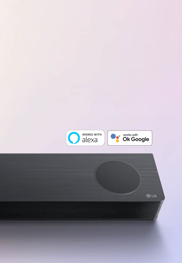 LG horizontalusis garsiakalbis ?Sound Bar“ pad?tas ant ?em?s, ant horizontaliojo garsiakalbio de?iniojo kampo pavaizduotas LG logotipas. Ant horizontaliojo garsiakalbio u?d?tas ?Alexa“ logotipas ir OK GOOGLE  logotipas.
