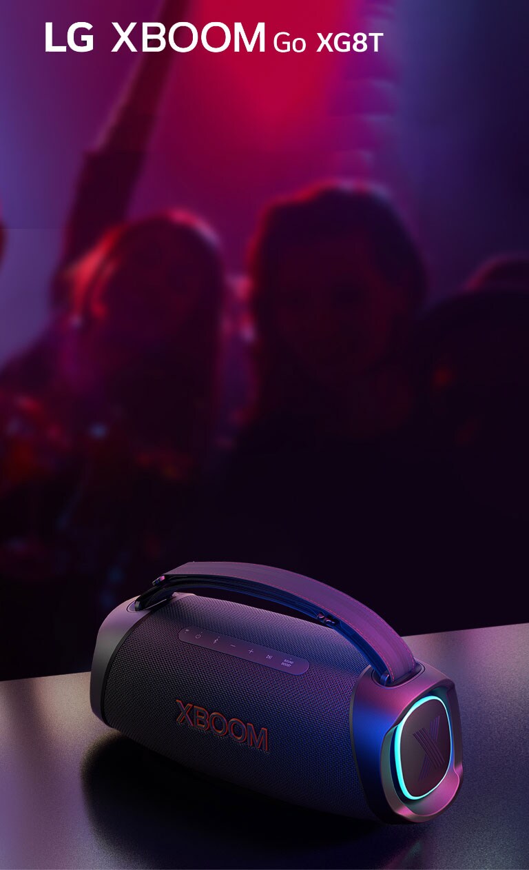 LG XBOOM Go XG8T pad?tas ant metalinio stalo su ?jungtu oran?iniu ap?vietimu. U? stalo ?mon?s m?gaujasi muzika.